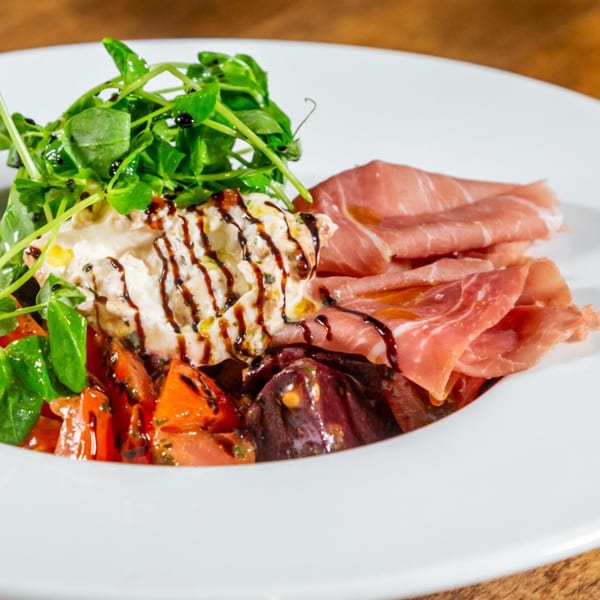 A white plate holds a salad with prosciutto, burrata, tomatoes, and greens drizzled with balsamic glaze.