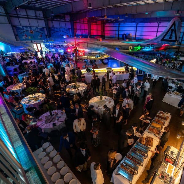 A large event with many people, buffet tables, and a vintage airplane under colorful lighting.