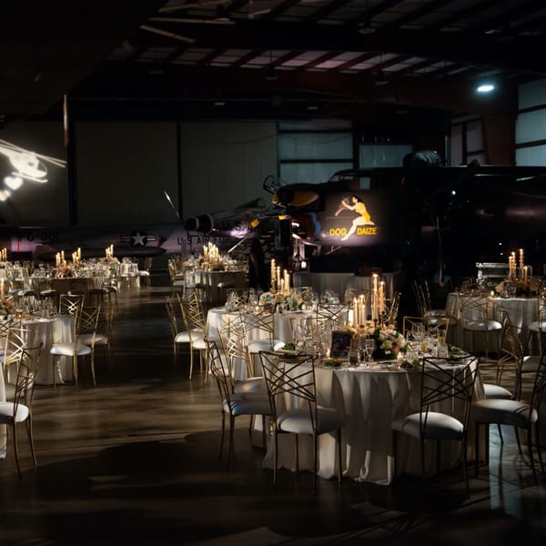 Elegant dining tables with candles and floral centerpieces set in a dimly lit hangar with airplanes.