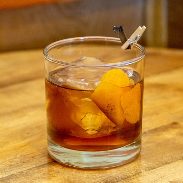 An Old Fashioned cocktail with a large ice cube, orange peel, and a small clothespin garnish.