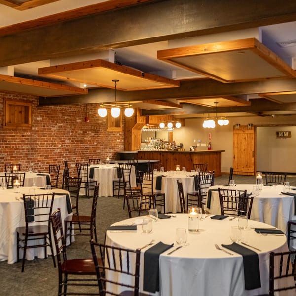 A large dining room with round tables set with white tablecloths, dark napkins, and candles.