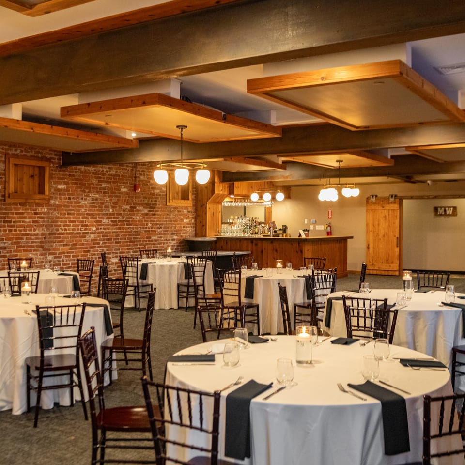 A large dining room with round tables set with white tablecloths, dark napkins, and candles.