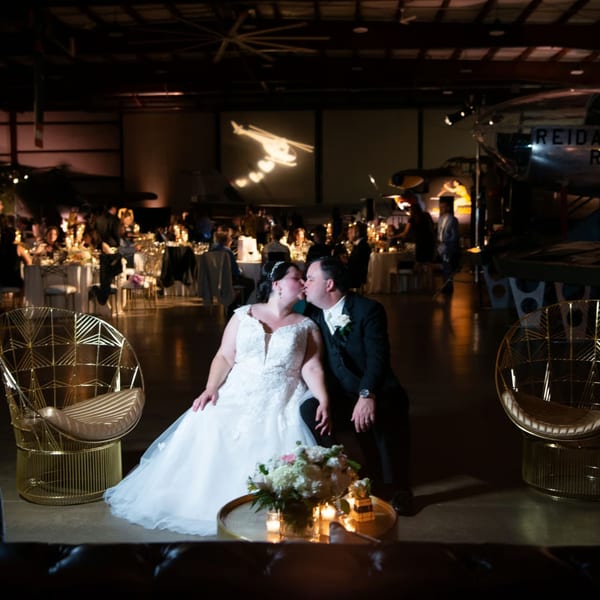 A bride and groom kiss in a dimly lit event space with tables and airplanes in the background.