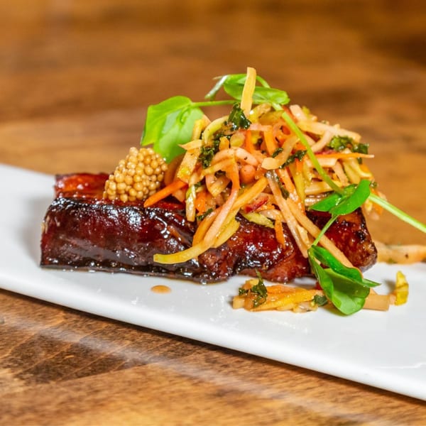 Glazed pork belly with pickled mustard seeds and a fresh vegetable slaw on a white plate.