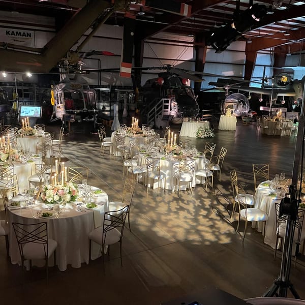 Elegant dining tables with white tablecloths and gold chairs set up in a large hangar with helicopters.