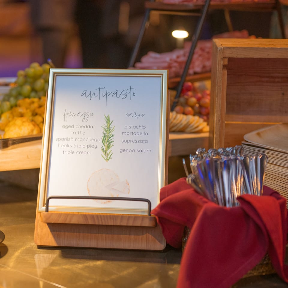 An antipasto sign lists cheeses and meats, with cutlery and food platters in the background.