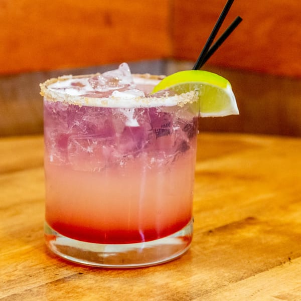 A vibrant pink and purple cocktail with ice, a lime wedge, and two black straws.