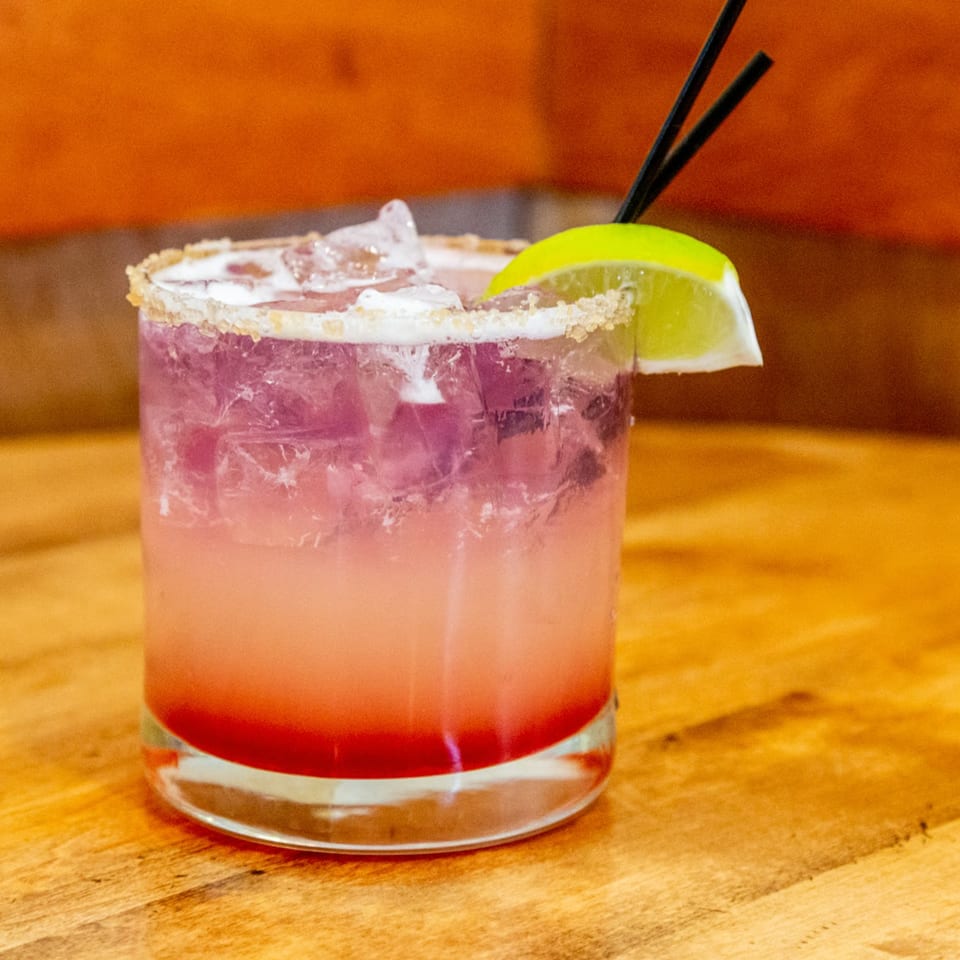 A vibrant pink and purple cocktail with ice, a lime wedge, and two black straws.