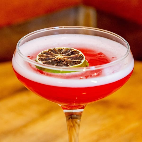 A vibrant red cocktail with a frothy white top, garnished with a dried lime slice.
